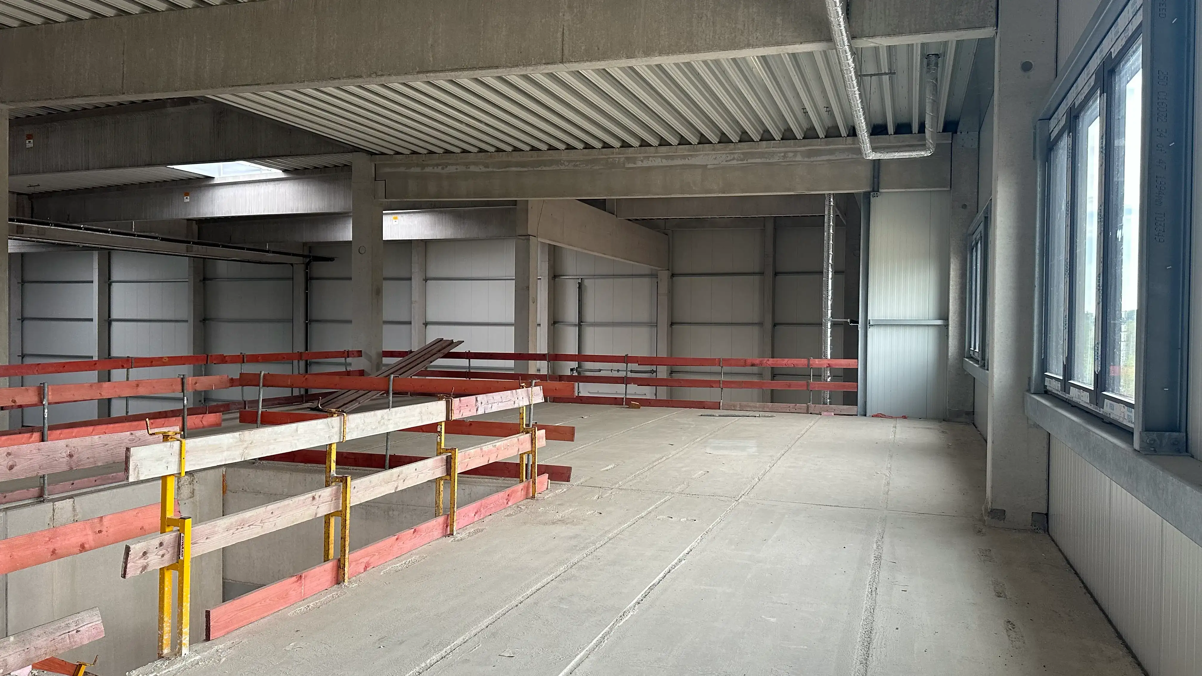 Baustelle: Rohbau der Firmenzentrale mit Betonstützen, Trapezblechdecke, rot-weißer Absturzsicherung an der Treppenöffnung und Fensterband.