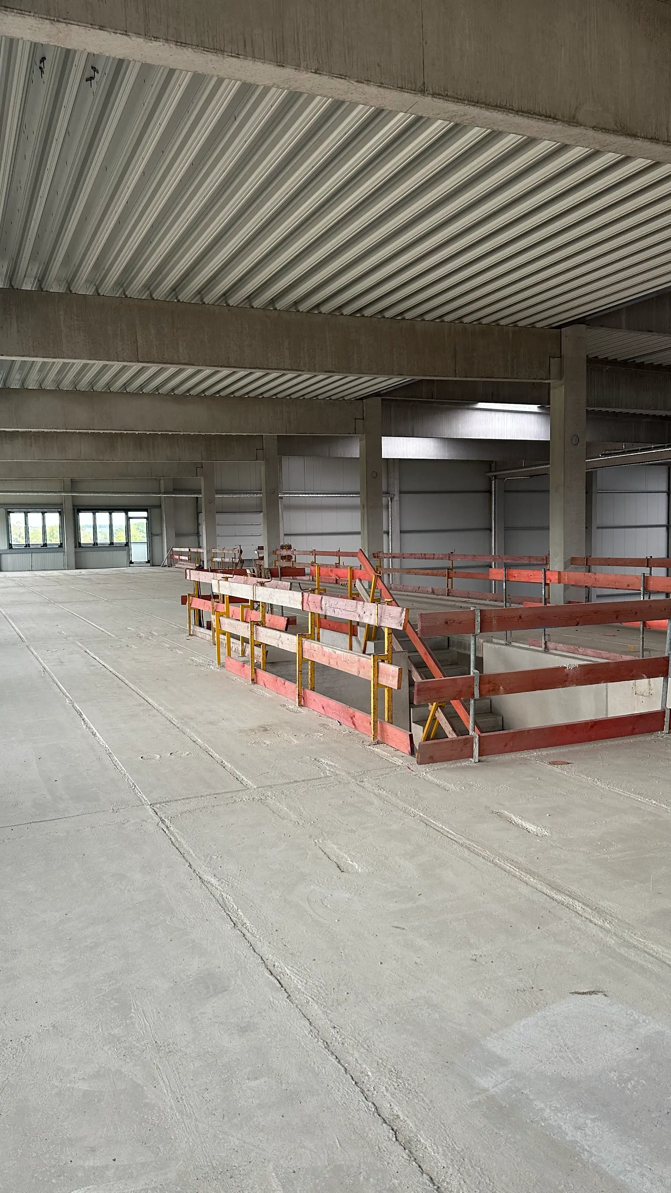 Baustelle: offene Rohbaufläche mit Betonboden, Trapezblechdecke und abgesicherter Treppenöffnung, im Hintergrund Fenster.
