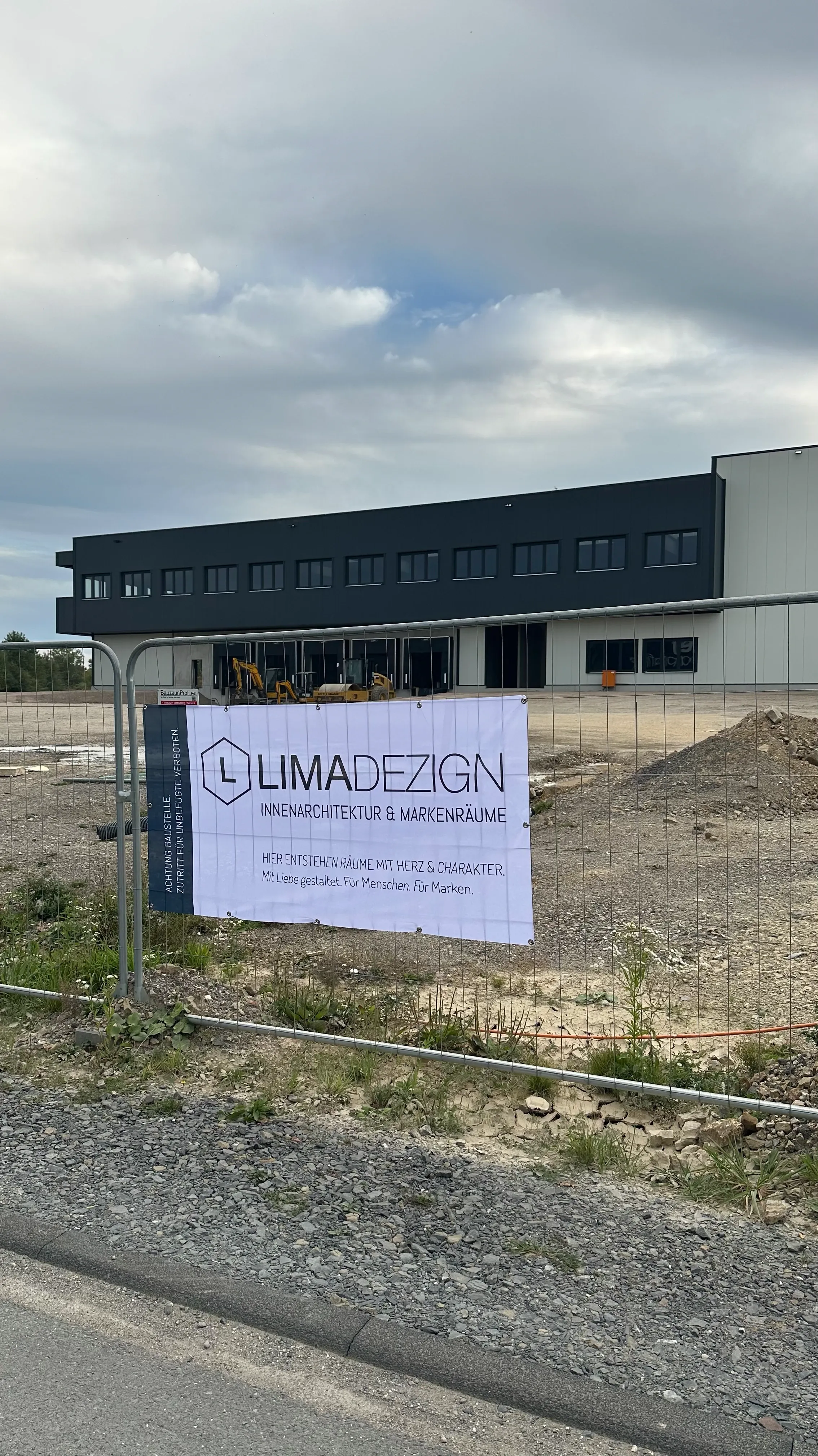 Baustelle von außen: Neubau der Firmenzentrale hinter einem Bauzaun mit limadezign-Banner „Hier entstehen Räume mit Herz & Charakter“.