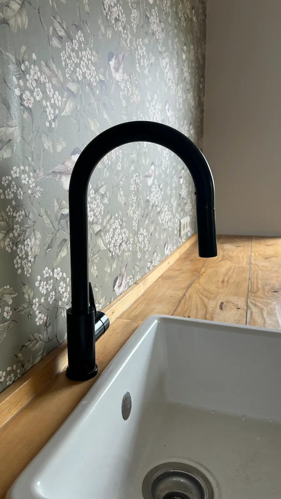 Wohnhaus K6 Detail: schwarze Armatur an einer weißen Keramikspüle mit Holzarbeitsplatte vor Mustertapete.